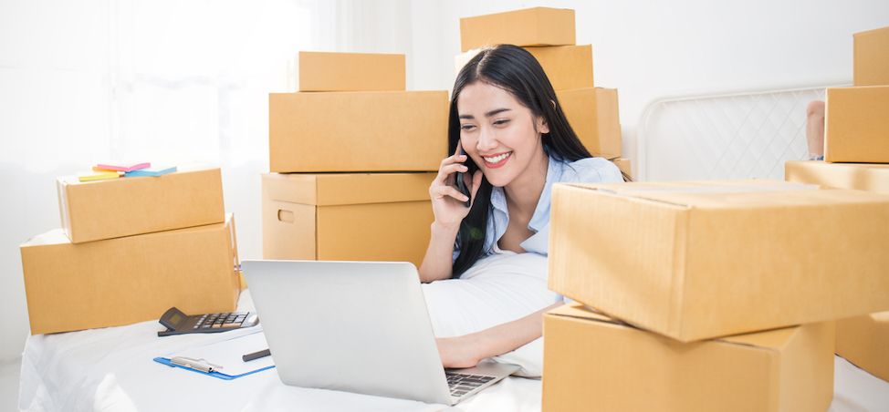 Young business woman sitting at desk in front of laptop on phone around boxes 