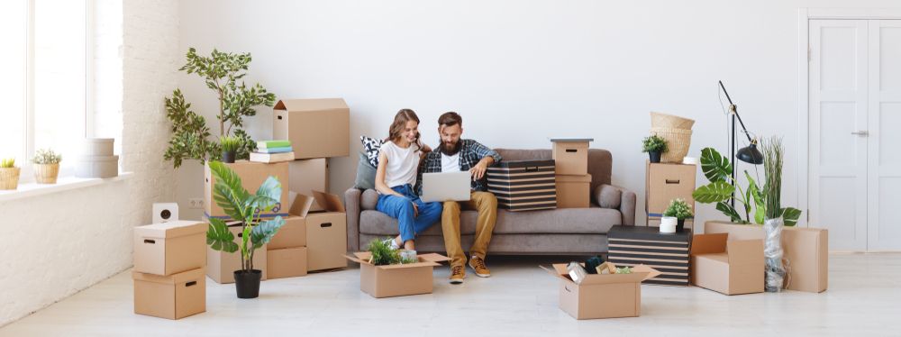 young couple sitting on couch surrounded by moving boxes looking at laptop