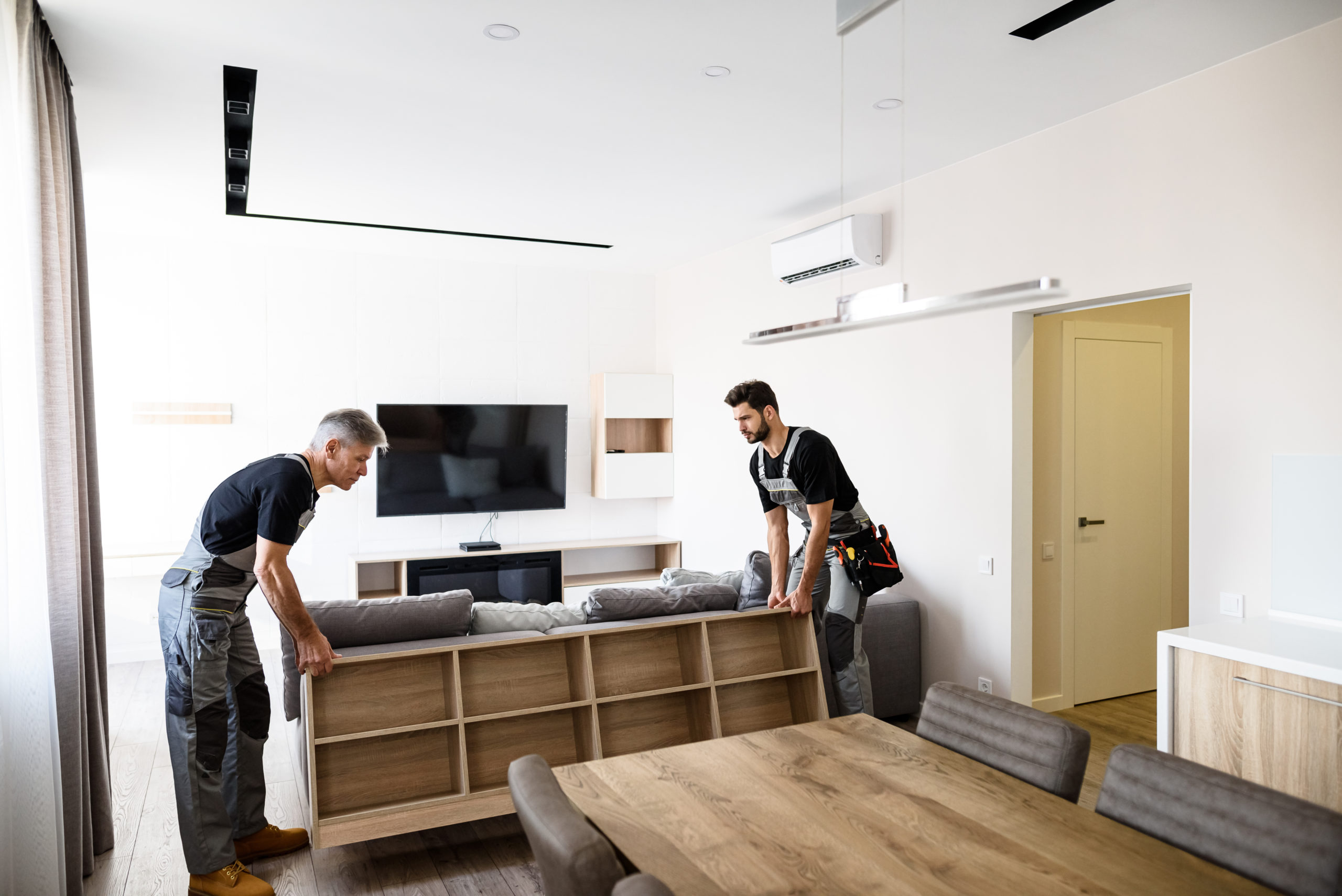 Full length shot of two contractor employees, young and aged worker in uniform moving personal belongings, furniture inside apartment. Moving service concept. Selective focus. Horizontal shot