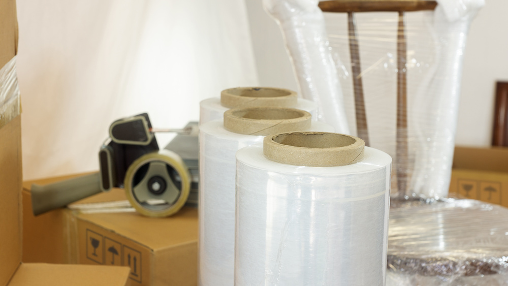Approach to plastic rolls for packing in the foreground with a plastic wrapped chair, cardboard boxes and adhesive tape in the background