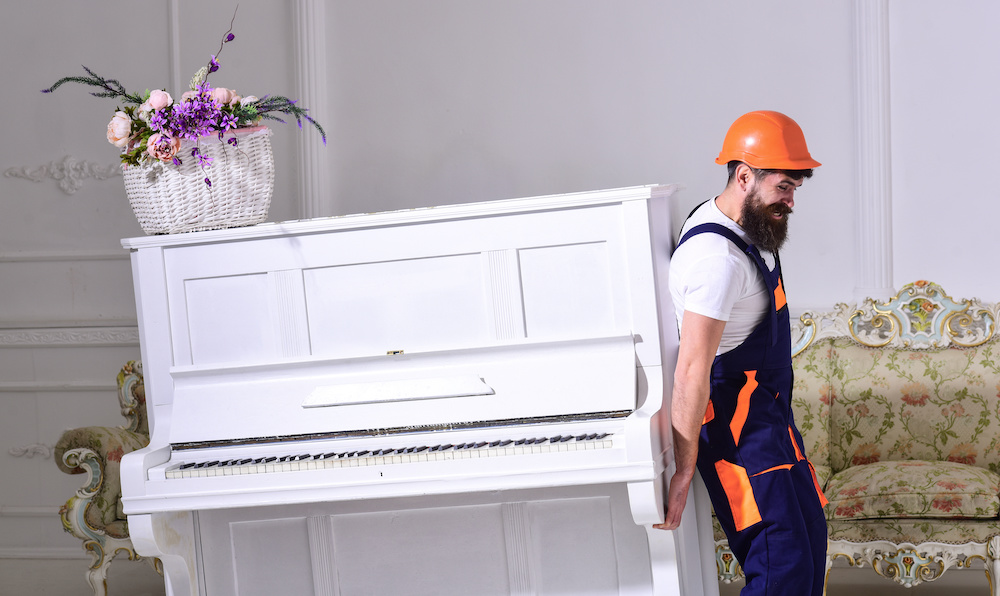 Loader moves piano instrument. Man with beard, worker in overalls and helmet lifts up piano, white background. Courier delivers furniture in case of move out, relocation. Delivery service concept.