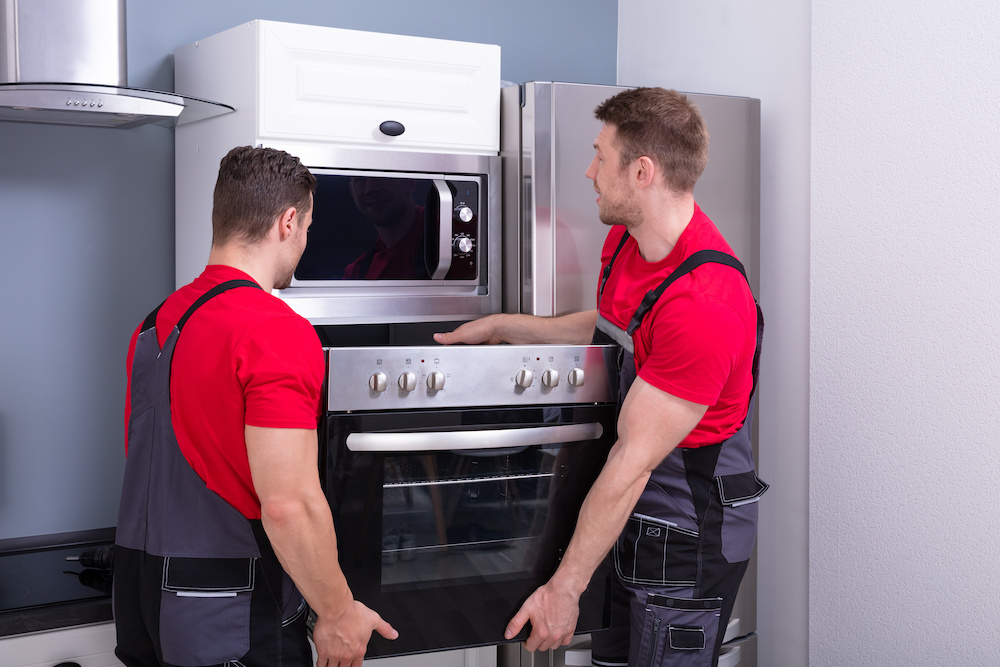 Side View Of Two Young Male Worker Placing Modern Oven Inside The Cabinet In Kitchen