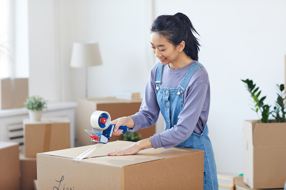 Portrait of smiling Asian woman packing cardboard boxes with tape dispenser while st moving out to new house, copy space