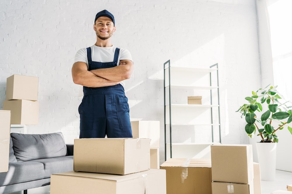 cheerful mover in uniform and cap standing with crossed arms near boxes in apartment