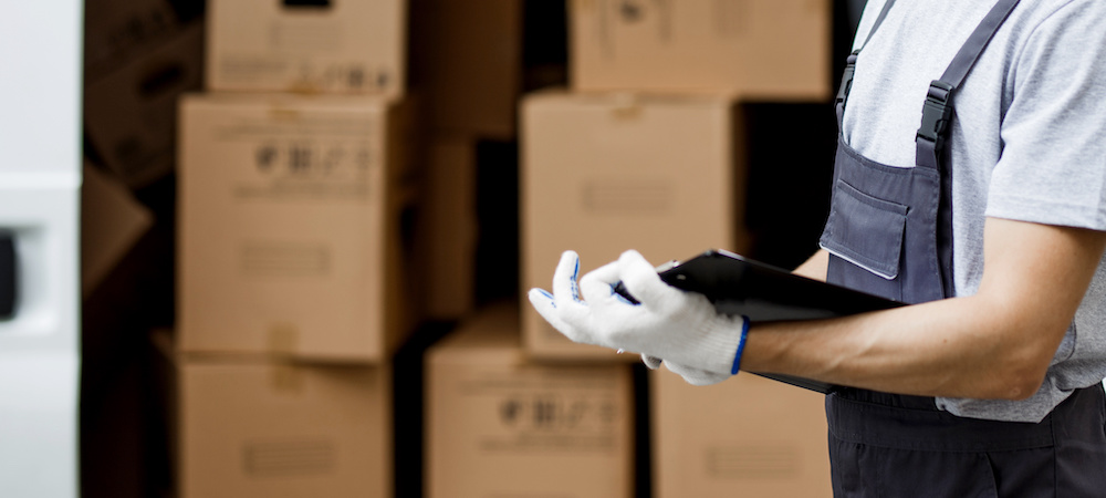 A young handsome smiling worker wearing uniform is standing next to the van full of boxes holding a clipboard in his hands. House move, mover service