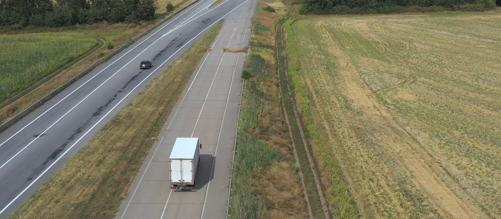 Aerial view of truck with cargo trailer driving on straight road and transporting goods. Camera following to delivery lorry moving across country highway. Flying over traffic in rural.
